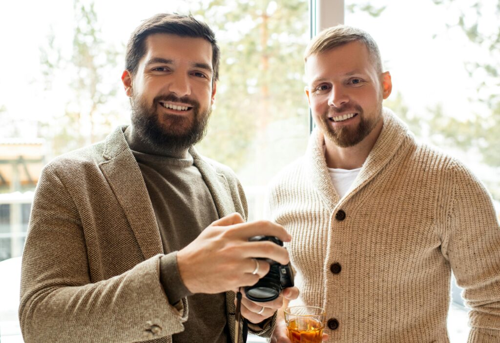 Two smiling men indoors, one holding a camera and the other a drink, in a relaxed setting.