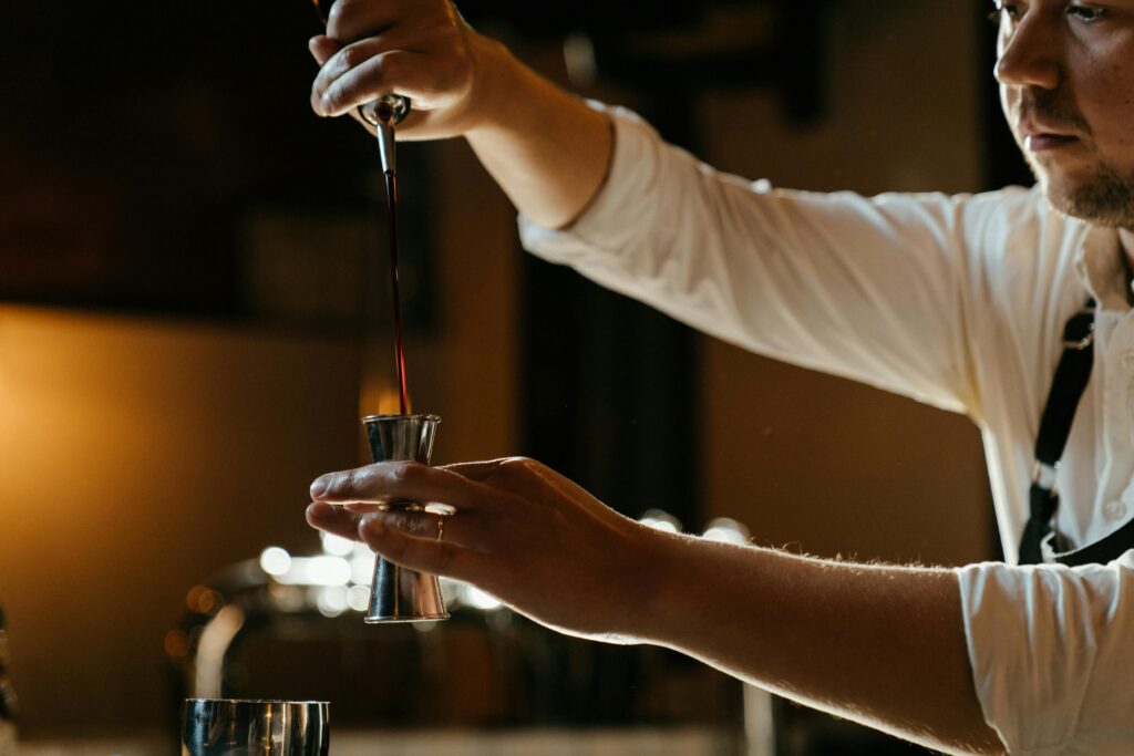 A bartender in a uniform expertly measures spirits with a jigger in a dimly lit indoor bar setting.